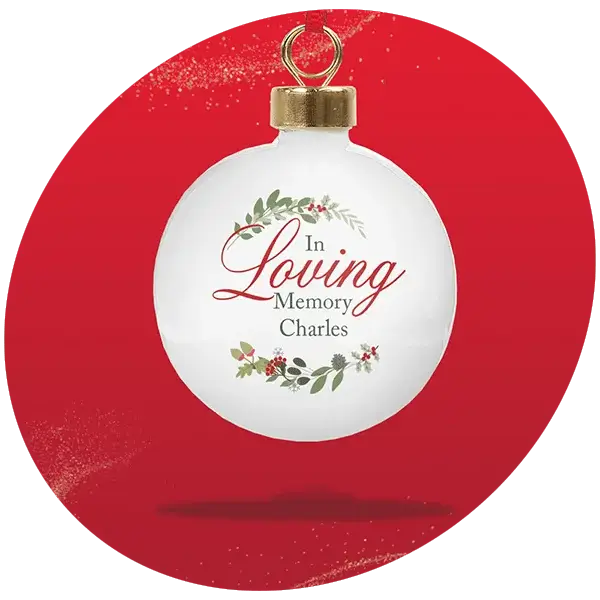 White Christmas bauble with gold cap and hanging loop, featuring the text “In Loving Memory Charles” surrounded by festive greenery and red berries, displayed against a bright red background.