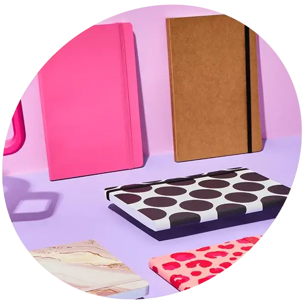 A collection of colorful notebooks displayed on a pink and purple surface, including a plain pink notebook, a brown kraft notebook with a black elastic band, a white notebook with large black polka dots, and two patterned notebooks featuring marble and pink leopard designs.