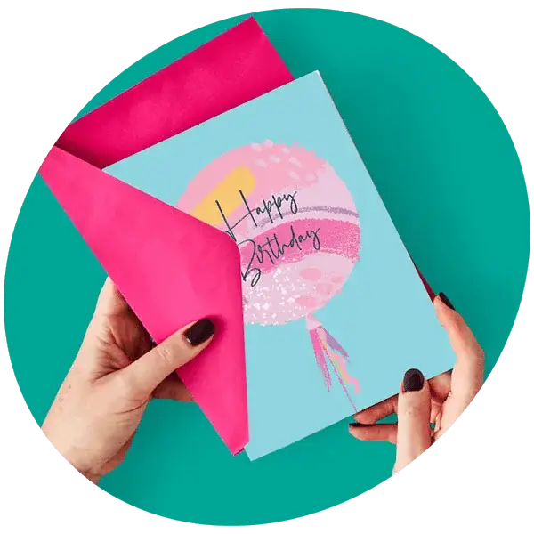 Hands holding a colourful Happy Birthday card with a pink envelope against a teal background.