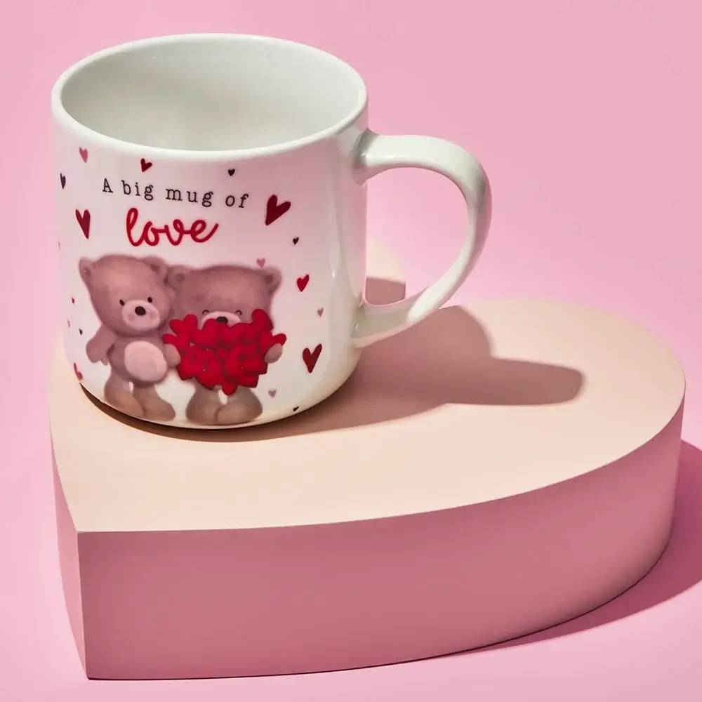 White ceramic mug with two teddy bears holding a red heart bouquet, surrounded by small red hearts and the text A big mug of love, placed on a light pink heart-shaped platform against a pink background.