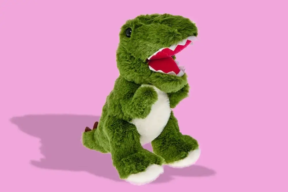 Green plush dinosaur toy with white belly and brown spikes, shown against a pink background. Green plush dinosaur toy with white belly and brown spikes, shown against a pink background.