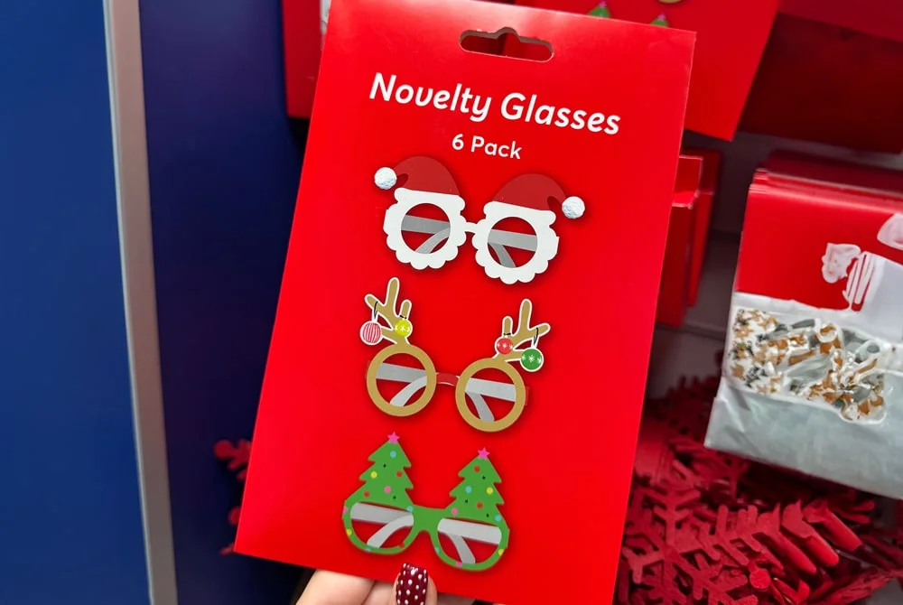A hand holding a red package labeled Novelty Glasses containing six festive glasses designs, including Santa hats, reindeer antlers, and Christmas trees, displayed in a store aisle with other holiday items in the background. A hand holding a red package labeled Novelty Glasses containing six festive glasses designs, including Santa hats, reindeer antlers, and Christmas trees, displayed in a store aisle with other holiday items in the background.