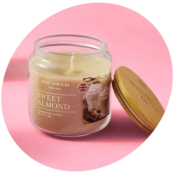 A sweet almond scented candle in a clear glass jar with a wooden lid resting beside it on a pink background.