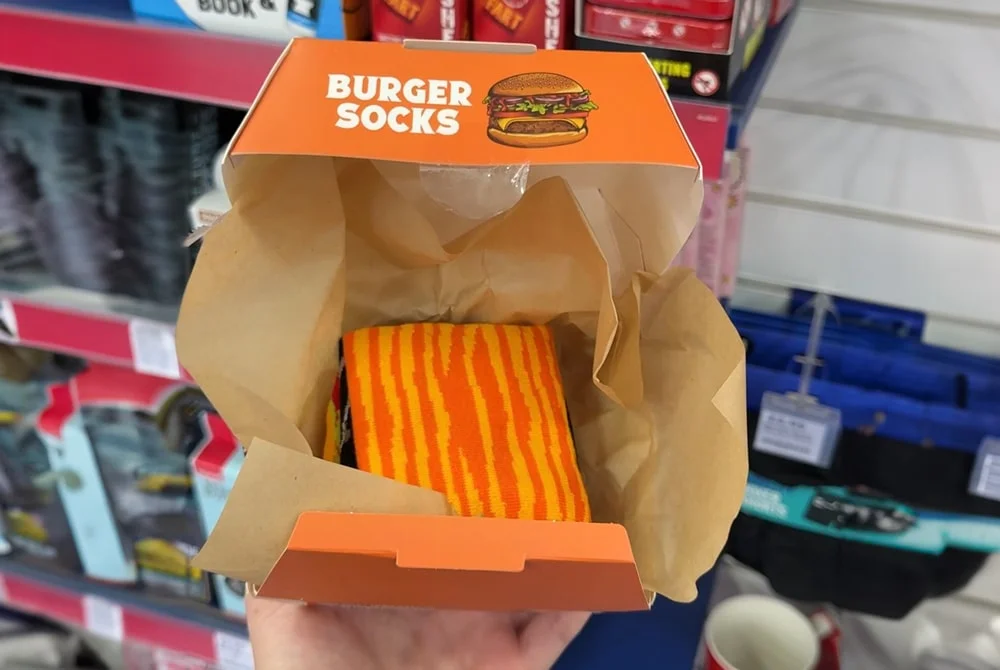 A novelty gift box labeled “Burger Socks” containing a pair of orange and yellow striped socks, designed to resemble a burger, displayed in a store setting. A novelty gift box labeled “Burger Socks” containing a pair of orange and yellow striped socks, designed to resemble a burger, displayed in a store setting.