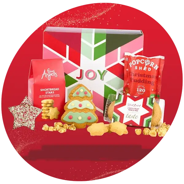 Festive gift hamper with a colorful box labeled Joy, surrounded by Christmas treats including shortbread stars, iced tree-shaped biscuits, popcorn, and mince pies, displayed against a red background with a glitter effect.