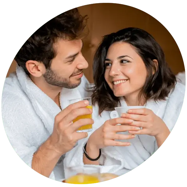 Two people wearing white bathrobes sit close together, holding drinks—one with a glass of orange juice and the other with a white cup—on a wooden surface with a breakfast tray in front of them.