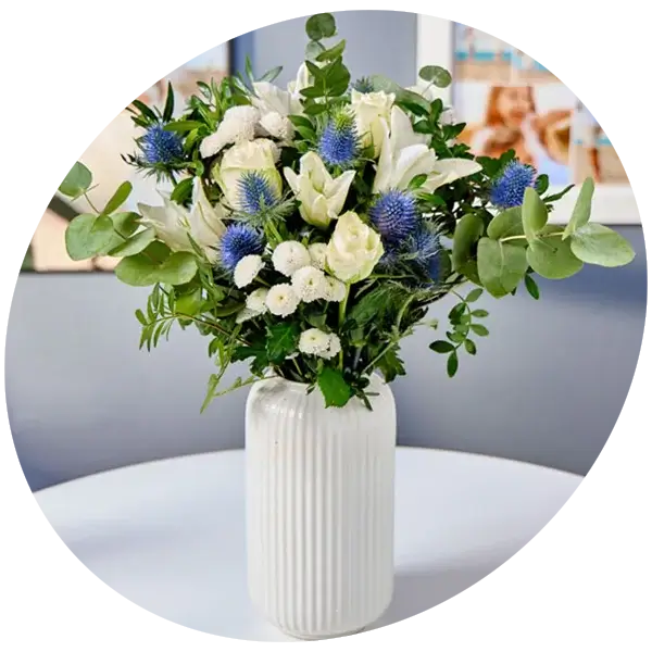 A bouquet of white and blue flowers with green foliage arranged in a tall ribbed white vase on a round table.