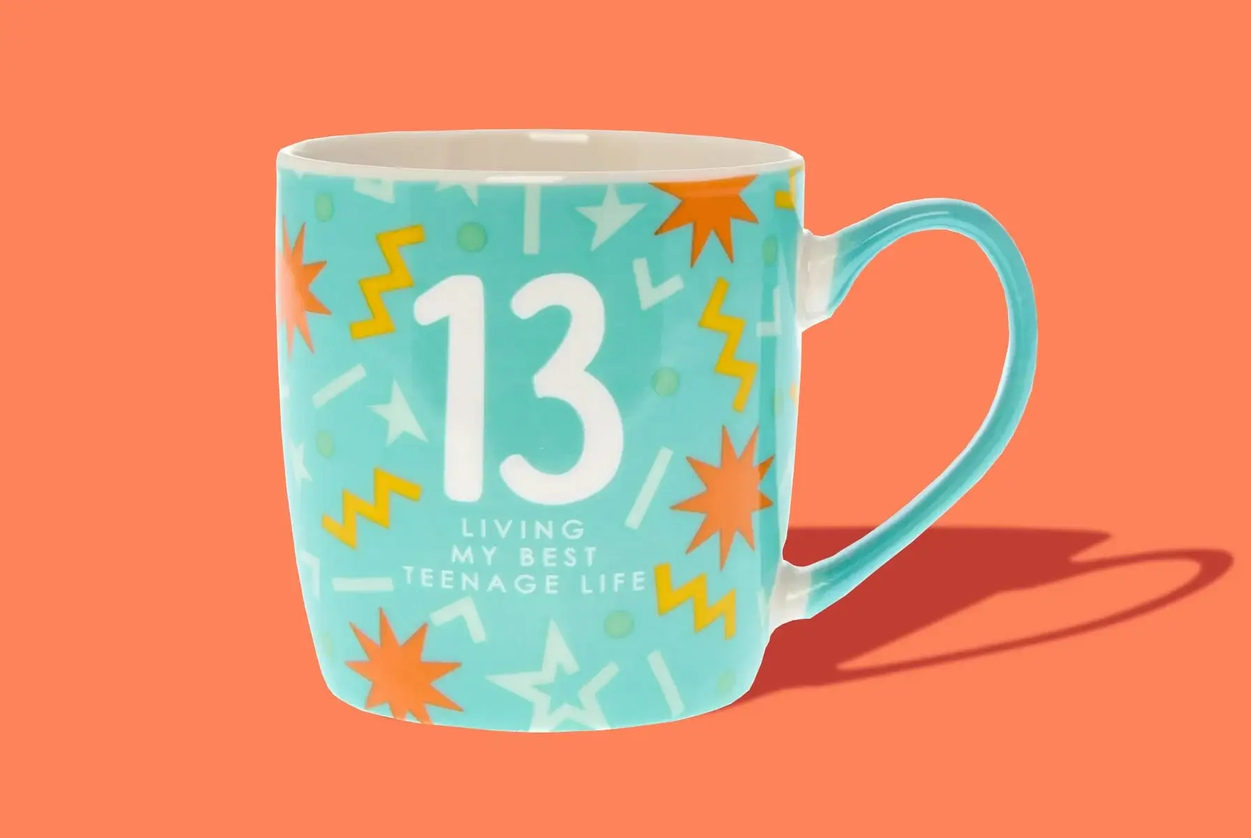 'A turquoise mug with the number 13 in large white font, surrounded by colorful starbursts and zigzag patterns. Below the number, the text reads: LIVING MY BEST TEENAGE LIFE.' 'A turquoise mug with the number 13 in large white font, surrounded by colorful starbursts and zigzag patterns. Below the number, the text reads: LIVING MY BEST TEENAGE LIFE.'