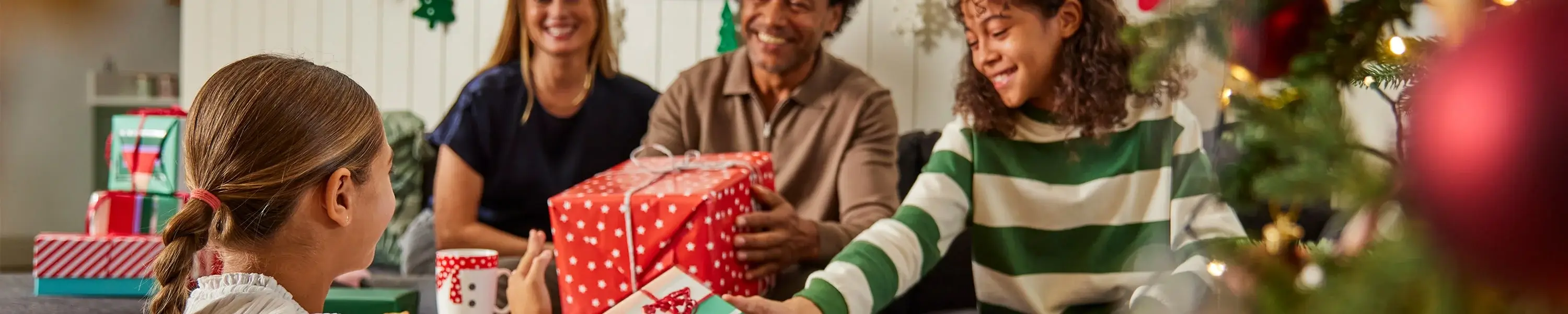 A festive living room scene with a decorated Christmas tree in the foreground and several wrapped presents on the floor. People are sitting together exchanging gifts, including a large red box with white snowflake patterns. The background features holiday decorations on the wall. A festive living room scene with a decorated Christmas tree in the foreground and several wrapped presents on the floor. People are sitting together exchanging gifts, including a large red box with white snowflake patterns. The background features holiday decorations on the wall.