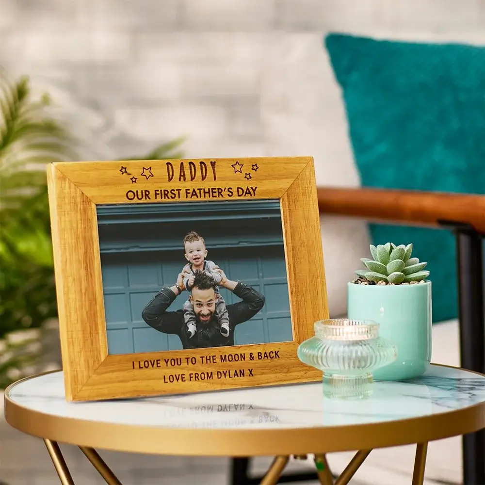 A wooden picture frame with the text 'DADDY' at the top, 'OUR FIRST FATHER'S DAY' in the middle, and 'I LOVE YOU TO THE MOON & BACK LOVE FROM DYLAN X' at the bottom. The frame contains a photo of an adult holding a child on their shoulders. The frame is placed on a round table with a small potted plant and two glass candle holders.