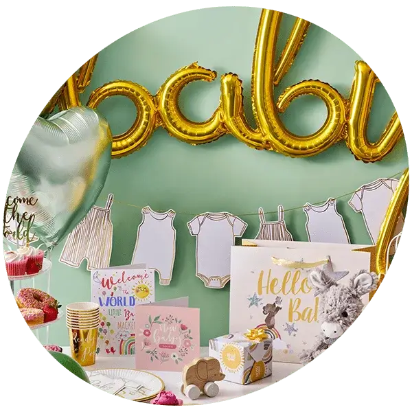 A baby shower setup with a green background and gold balloon letters spelling 'baby'. Below the balloons, there are paper cutouts of baby onesies hanging on a string. The table is decorated with various items including a silver heart-shaped balloon, cupcakes, greeting cards with messages like 'Welcome to the World Little One' and 'Hello Baby', gold cups, plates, and napkins. There is also a small wooden toy car and plush animal.