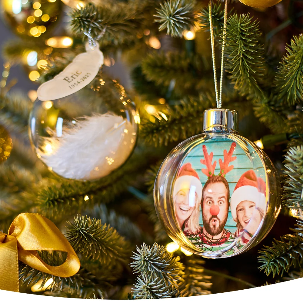 Close-up of a Christmas tree with two clear baubles: one with a white feather and the name 'Eric' with dates, and another showing a photo of three people in festive hats and accessories.