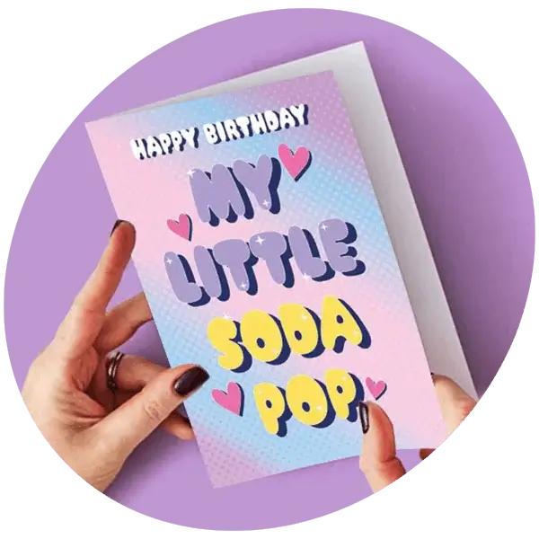 A person holding a colourful birthday card with the text Happy Birthday My Little Soda Pop against a purple background.