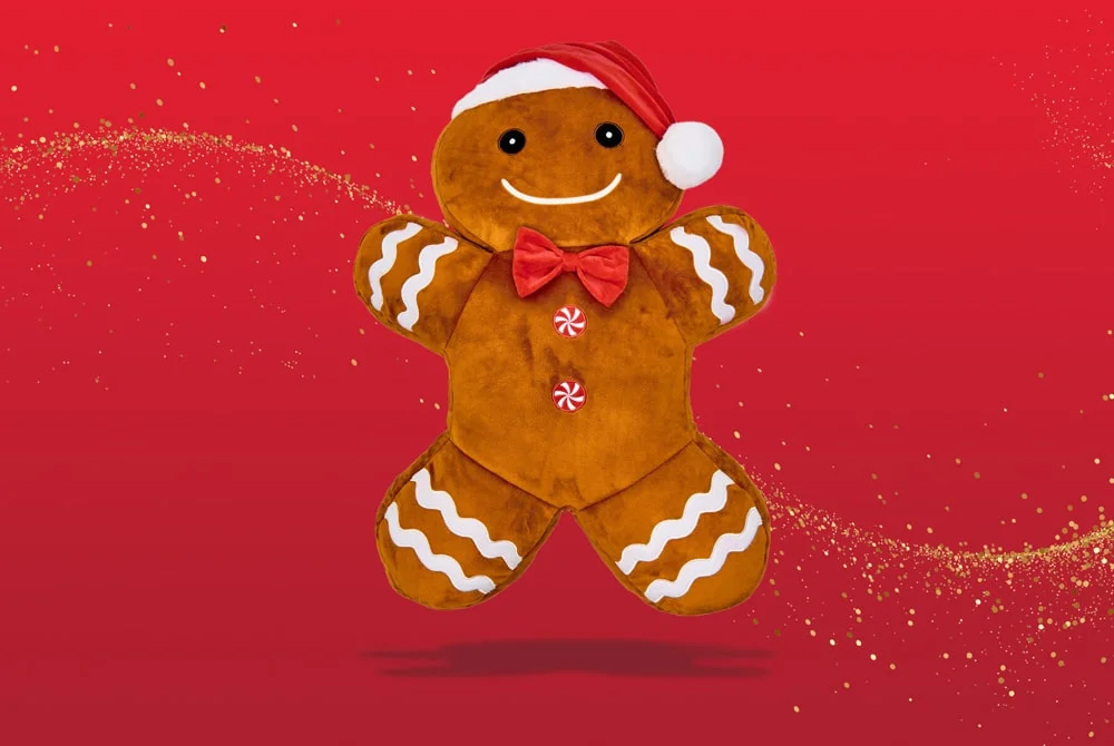 A plush gingerbread figure wearing a red Santa hat and bow tie, decorated with white icing details, set against a bright red festive background with gold sparkles. A plush gingerbread figure wearing a red Santa hat and bow tie, decorated with white icing details, set against a bright red festive background with gold sparkles.