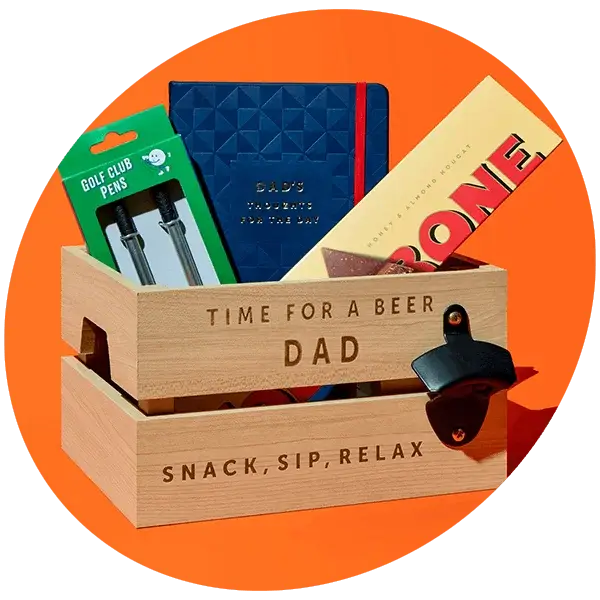 A wooden crate engraved with Time for a Beer Dad and Snack Sip Relax, filled with Father’s Day gifts including a blue notebook, Toblerone chocolate and golf club pens, set against a bright orange background.