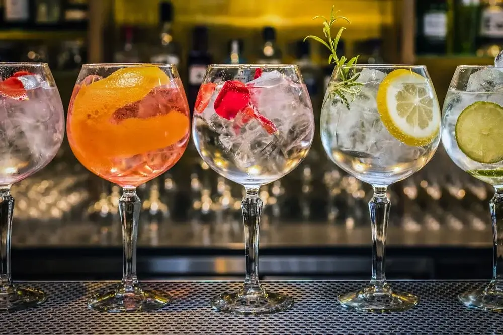 Five large gin glasses filled with ice and garnished with colorful fruits and herbs, including strawberries, orange peel, lemon slices, cucumber, and rosemary, displayed on a bar counter with bottles in the background. Five large gin glasses filled with ice and garnished with colorful fruits and herbs, including strawberries, orange peel, lemon slices, cucumber, and rosemary, displayed on a bar counter with bottles in the background.