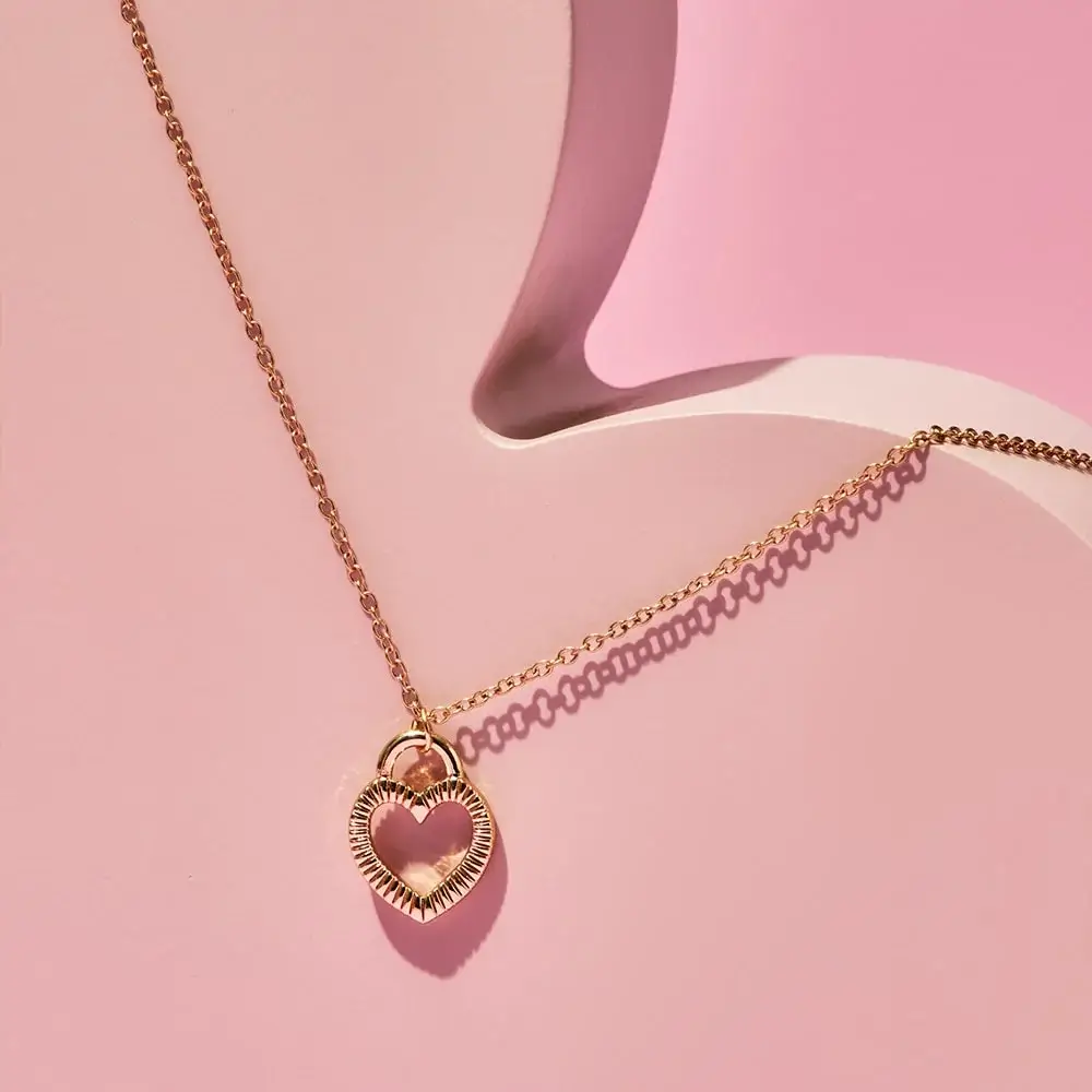 Red and gold artificial rose displayed under a clear glass dome on a round base, surrounded by pink and red paper hearts on a pink background.