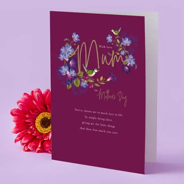 A deep purple Mother’s Day card decorated with colourful flowers and small birds, displayed upright beside a bright pink flower on a soft lilac background.