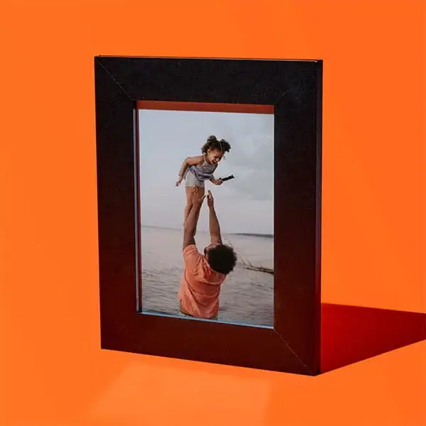 Black photo frame on an orange background showing a person lifting a child above their head by the sea