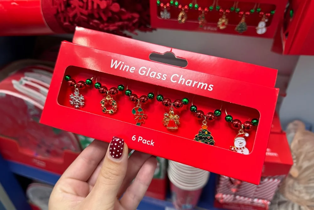A hand holding a red box labeled Wine Glass Charms containing six festive charms with designs like snowflakes, wreaths, Christmas trees, and Santa, displayed in a store aisle with other holiday items in the background. A hand holding a red box labeled Wine Glass Charms containing six festive charms with designs like snowflakes, wreaths, Christmas trees, and Santa, displayed in a store aisle with other holiday items in the background.