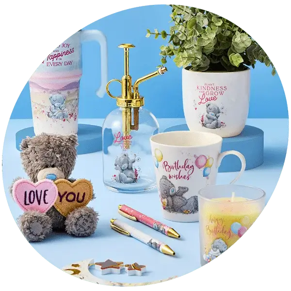 A collection of cute and colorful items arranged on a blue surface. Items include mugs with messages like 'Enjoy Happiness Every Day' and 'Birthday wishes', a plush teddy bear holding hearts that say 'LOVE YOU', a candle labeled 'Happy Birthday', a spray bottle, pens with teddy bear designs, and star-shaped decorations.