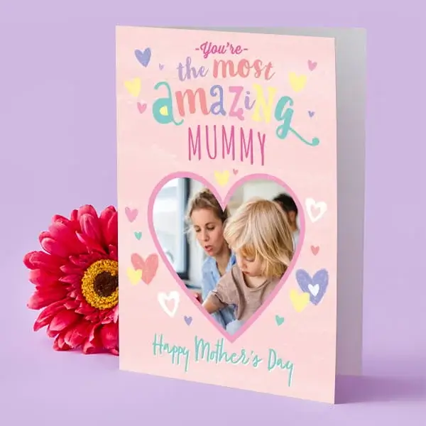 A pink Mother’s Day card decorated with colourful hearts and a heart-shaped photo window, displayed upright beside a bright pink flower.