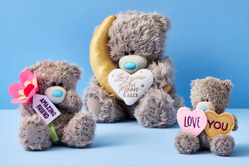 Three plush teddy bears with blue noses are displayed against a light blue background. The largest bear holds a golden crescent moon and a white heart with the text 'Love you to the Moon & Back'. The medium-sized bear holds a pink flower with the text 'AMAZING FRIEND'. The smallest bear holds two hearts, one pink and one yellow, with the text 'LOVE' on the pink heart and 'YOU' on the yellow heart.