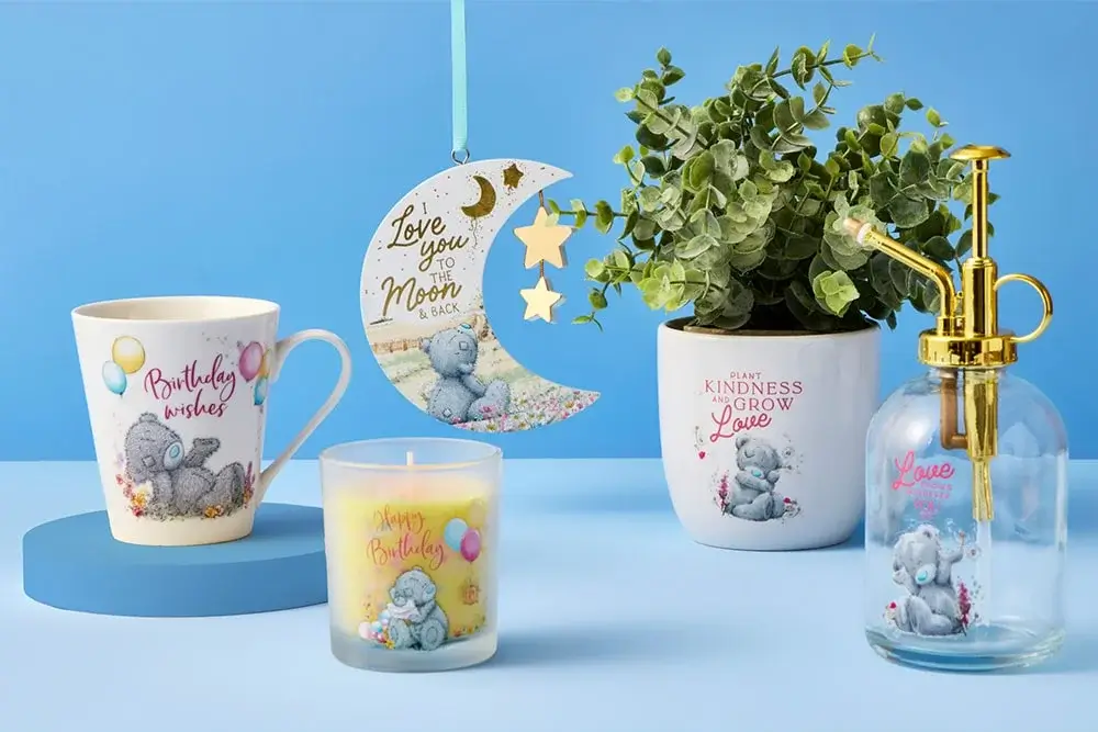 The image shows a collection of decorative items on a light blue surface with a light blue background. There is a white mug with the text 'Birthday wishes' and an illustration of a gray teddy bear holding balloons. Next to it, there is a candle in a glass holder with the text 'Happy Birthday' and the same teddy bear illustration. Hanging above these items is a crescent moon-shaped decoration with the text 'Love you to the Moon & back' and another teddy bear illustration. To the right, there is a white plant pot with green foliage and the text 'Plant kindness grow love' along with another teddy bear illustration. Finally, there is a clear glass soap dispenser with gold pump hardware and pink text that reads 'Love grows here,' also featuring the same teddy bear.