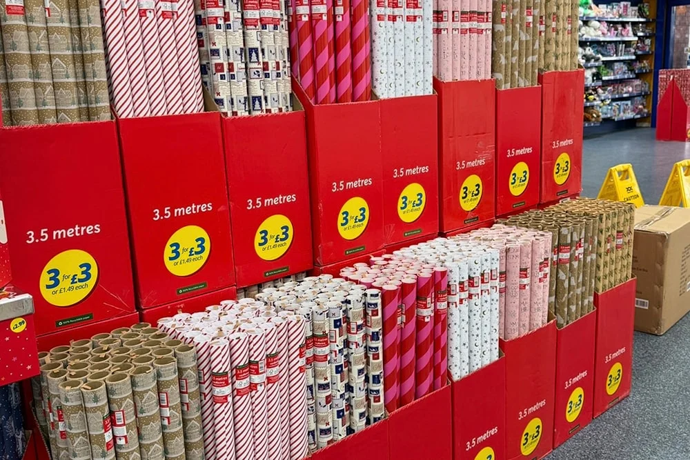 Store display of assorted Christmas wrapping paper rolls in red boxes labeled 3.5 metres, featuring festive patterns like stripes, snowflakes, and reindeer. Store display of assorted Christmas wrapping paper rolls in red boxes labeled 3.5 metres, featuring festive patterns like stripes, snowflakes, and reindeer.