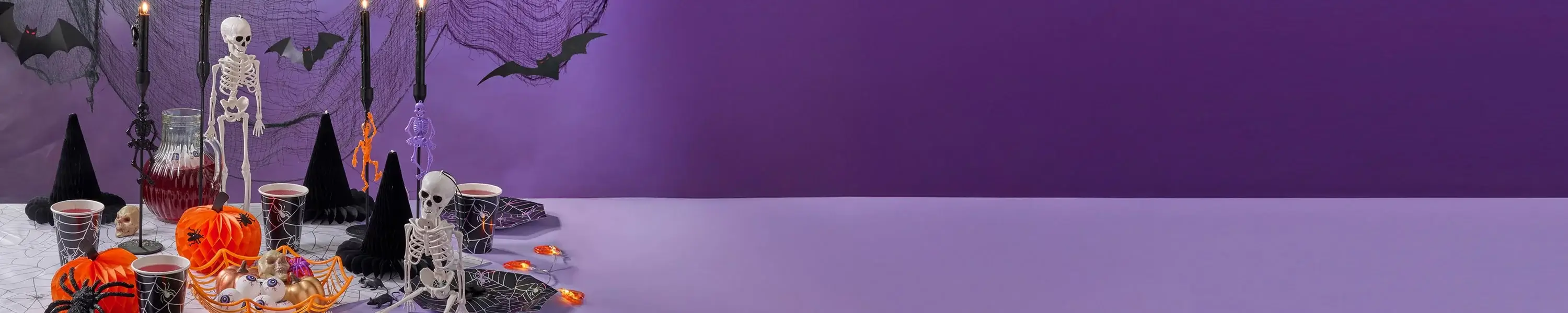 Halloween table setup with skeletons, black witch hats, orange pumpkins, spider webs, and candles on a purple background. Halloween table setup with skeletons, black witch hats, orange pumpkins, spider webs, and candles on a purple background.