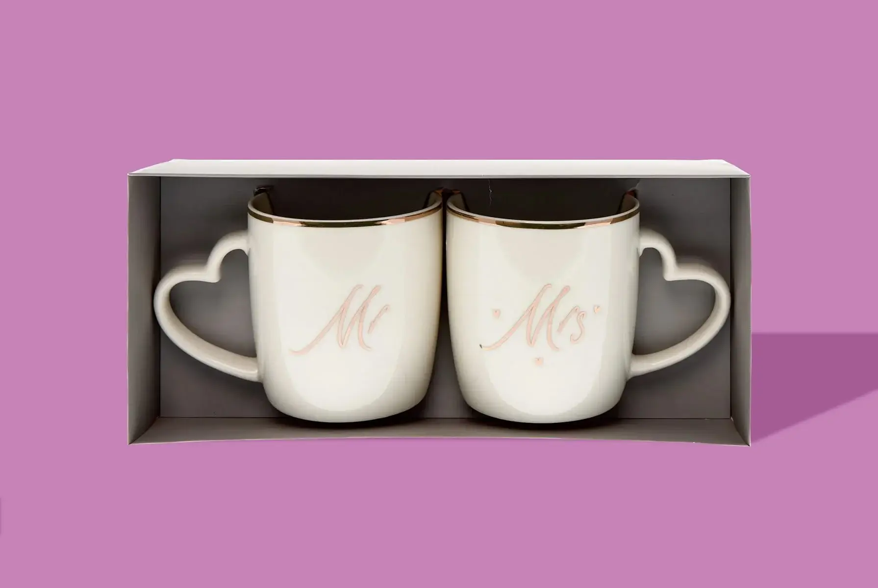 'A set of two white ceramic mugs with heart-shaped handles, displayed in a grey box against a pink background. The left mug has the word 'Mr' written in cursive pink letters, and the right mug has the word 'Mrs' written in cursive pink letters.' 'A set of two white ceramic mugs with heart-shaped handles, displayed in a grey box against a pink background. The left mug has the word 'Mr' written in cursive pink letters, and the right mug has the word 'Mrs' written in cursive pink letters.'