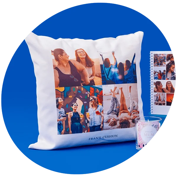 White cushion with a collage of six photos showing joyful moments, labeled Frans Cushion at the bottom, set against a solid blue background with a glass candle holder and a photo calendar nearby.