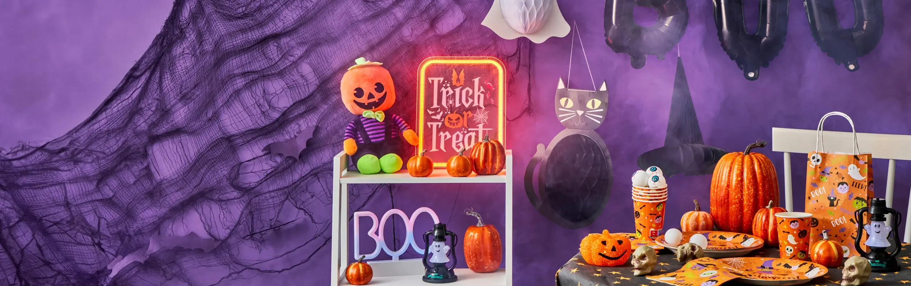Halloween-themed display with a purple backdrop, pumpkin plush toy, neon Trick or Treat sign, festive tableware, and hanging bat and cat decorations.