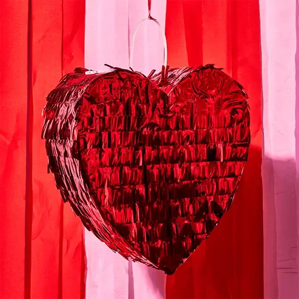 A shiny red heart‑shaped piñata hangs against a backdrop of red and pink streamers.
