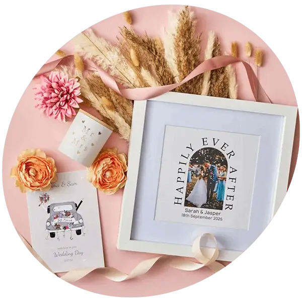 A wedding keepsake display on a pink background featuring a framed happily ever after photo print, decorative ribbons, dried flowers, and personalised cards.