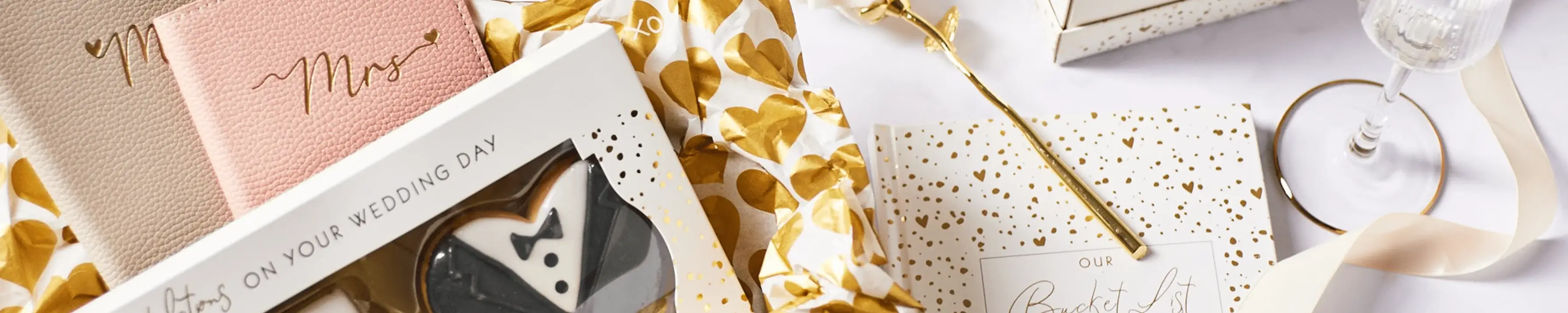 The image shows a collection of wedding-themed items arranged on a white surface. There are two greeting cards with the text 'Mr' and 'Mrs' written in gold script, a box labeled 'On Your Wedding Day' containing black and white cookies shaped like a tuxedo and wedding dress, a golden floral-patterned cloth, an open book titled 'Our Bucket List', and a wine glass with gold accents. The image shows a collection of wedding-themed items arranged on a white surface. There are two greeting cards with the text 'Mr' and 'Mrs' written in gold script, a box labeled 'On Your Wedding Day' containing black and white cookies shaped like a tuxedo and wedding dress, a golden floral-patterned cloth, an open book titled 'Our Bucket List', and a wine glass with gold accents.